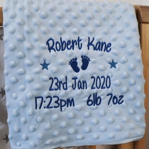 Op de afbeelding: Lichtblauwe babydeken met geborduurde tekst: "Robert Kane", "23rd Jan 2020", "17:23pm", "6lb 7oz" en twee sterren. De deken heeft een textuur.