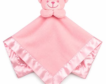 May include: A soft pink security blanket featuring a plush teddy bear head attached to the center. The square blanket is made of a fleece-like material and is finished with a smooth, shiny satin border along all four edges of the fabric.