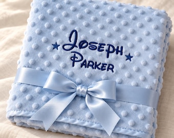 May include: A light blue dimpled minky blanket featuring the embroidered dark blue text Joseph Parker flanked by two small stars. The folded blanket is wrapped with a matching light blue satin ribbon tied into a decorative bow at the center.