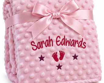 May include: A soft, light pink dimpled minky blanket featuring a satin ribbon tied in a bow. The blanket is embroidered with the name Sarah Edwards in dark pink thread, accompanied by a pair of small baby footprints and three dark pink stars.