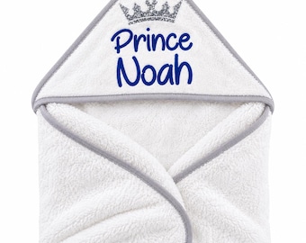 May include: A white hooded towel with grey trim featuring a silver glitter crown graphic and the text Prince Noah embroidered in blue script. The soft, plush fabric is folded to display the hood and the personalized name clearly on the front.