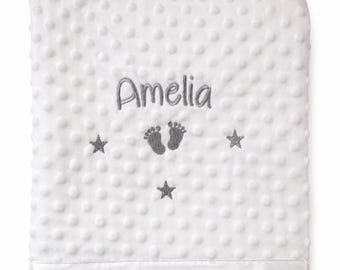 May include: A white dimpled minky blanket featuring the embroidered name Amelia in grey thread. The design includes a pair of grey baby footprints centered below the name, flanked by three grey embroidered stars arranged in a triangular pattern.