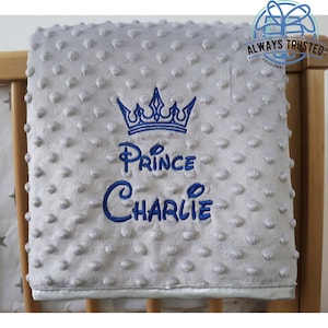 May include: A gray and white baby blanket with a blue embroidered crown, the words "Prince Charlie" and a white satin trim.