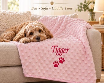 May include: A soft pink dimpled texture blanket featuring the embroidered name Tigger and two red paw prints. The text A COSY PLACE TO REST and Bed • Sofa • Cuddle Time is visible above a brown dog resting on the blanket atop a beige sofa.