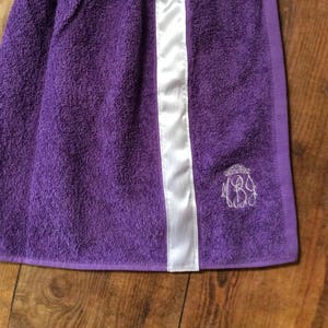 May include: A purple towel with a white satin stripe down the center. The towel has a monogram in the lower right corner. The towel is made of a soft, absorbent material.