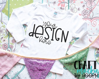 Puede incluir: Camiseta blanca de manga larga con el texto "your design here" sobre un fondo blanco. La camiseta está sobre una mesa con una guirnalda de lentejuelas rosa y verde.