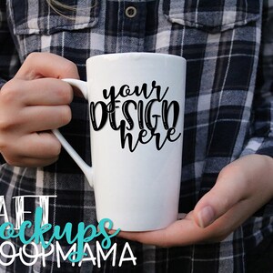May include: White ceramic mug with a black inscription that reads "your design here". The mug is being held by a person wearing a blue and black plaid shirt.