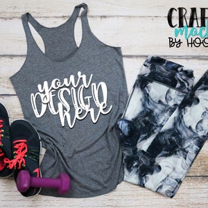 May include: A gray tank top with the text "your design here" in white, a pair of black and white leggings, a purple dumbbell, and two black running shoes with red laces on a wood background.