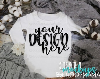 Puede incluir: Un body blanco de manga larga para bebé con un diseño negro que dice "your design here". El body está sobre una superficie de madera con una manta gris y bolas de algodón.