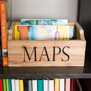 May include: A wooden box with the word "MAPS" printed on the front. The box is filled with folded maps. The box is sitting on a shelf with other books.