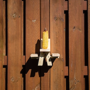 May include: A wooden corn feeder shaped like a miniature picnic table, holding an ear of yellow corn. The feeder is mounted on a brown wooden fence. The image is well-lit, with the corn and feeder as the focal point.