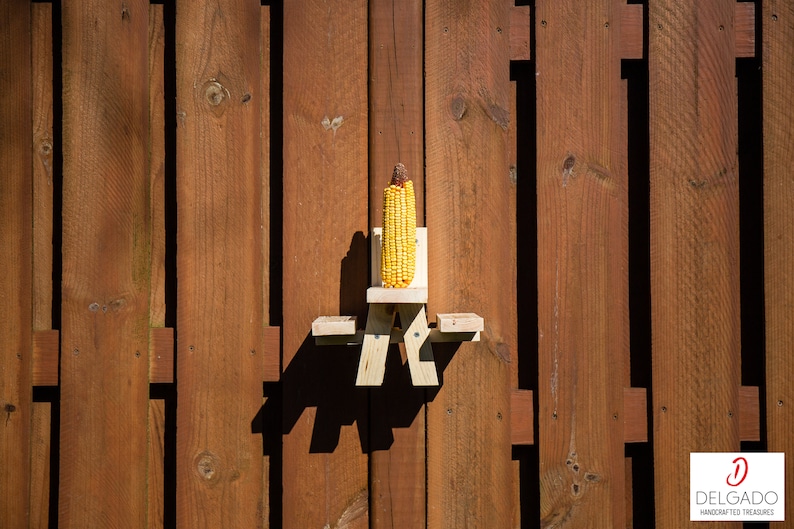 May include: A wooden corn feeder shaped like a miniature picnic table, holding an ear of yellow corn. The feeder is mounted on a brown wooden fence. The image is well-lit, with the corn and feeder as the focal point.
