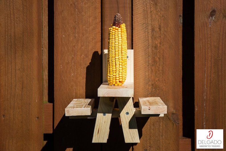 May include: A wooden corn feeder shaped like a picnic table, with a full ear of yellow corn. The feeder is mounted on a brown wooden fence. The corn has a brown, dried top. The feeder is made of light-colored wood.