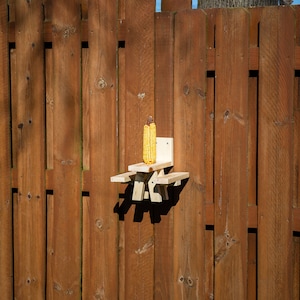 May include: A wooden squirrel feeder shaped like a picnic table, mounted on a brown wooden fence. The feeder holds an ear of yellow corn. The fence is made of vertical wooden planks.