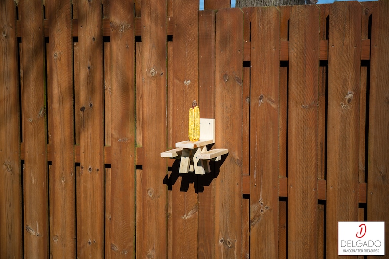 May include: A wooden squirrel feeder shaped like a picnic table, mounted on a brown wooden fence. The feeder holds an ear of yellow corn. The fence is made of vertical wooden planks.