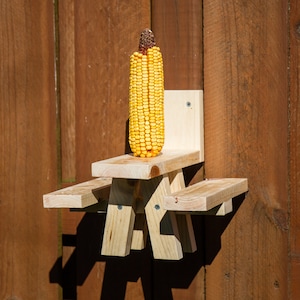 May include: A wooden squirrel feeder shaped like a picnic table, mounted on a wooden fence. A full ear of yellow corn sits on the table. The feeder is made of light-colored wood and is attached with screws.