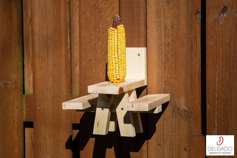 May include: A wooden squirrel feeder shaped like a picnic table, mounted on a wooden fence. A full ear of yellow corn sits on the table. The feeder is made of light-colored wood and is attached with screws.