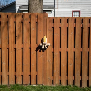 May include: A wooden fence with vertical planks stained in a warm brown hue. A small, handmade bird feeder is attached to the fence. The bird feeder is made of light-colored wood and has a yellow object inside. The Delgado logo is in the bottom right corner.