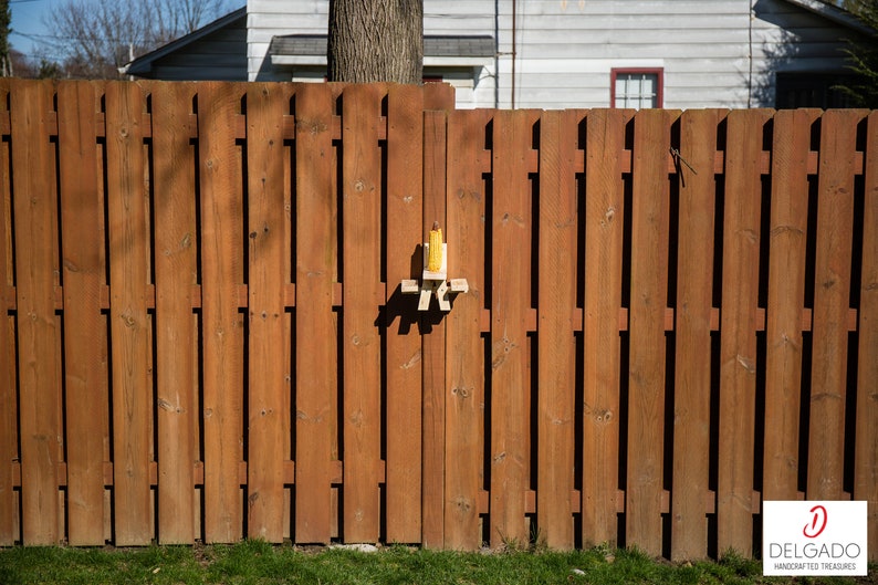 May include: A wooden fence with vertical planks stained in a warm brown hue. A small, handmade bird feeder is attached to the fence. The bird feeder is made of light-colored wood and has a yellow object inside. The Delgado logo is in the bottom right corner.
