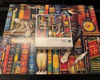 May include: A completed jigsaw puzzle and its box featuring a whimsical library scene with sleeping cats, books, an owl, and a clock. The puzzle box reads "750 pieces". The puzzle is a colorful illustration of a cozy reading nook.