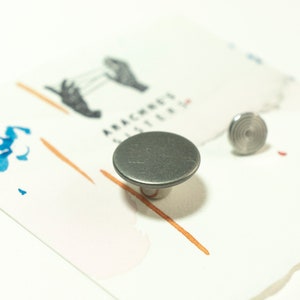 May include: Two silver metal buttons, one round and one with a spiral design, on a white surface with orange and blue paint splatters.