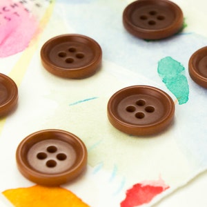 May include: Six brown buttons with four holes each, arranged in a row on a white surface with watercolor paint splatters.