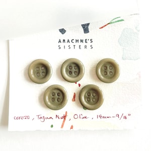 May include: Five olive-colored corozo buttons with four holes each, arranged on a white card. The card has the text "ARACHNE'S SISTERS" and handwritten details: "corozo, Tagua Nutt, Olive, 14mm-9/16".