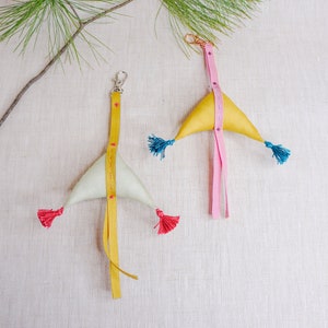 Op de afbeelding: Twee stoffen sleutelhangers in de vorm van driehoeken. Eén is geel met roze lint en blauwe kwastjes. De andere is wit met geel lint en rode kwastjes.