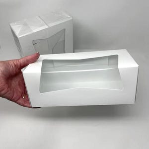 May include: White cardboard box with a clear plastic window. The box is empty and ready to be filled with a product.