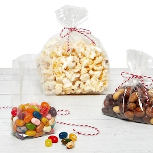 May include: Three clear cellophane bags filled with snacks. One bag contains popcorn, another jelly beans, and the third contains nuts. Each bag is tied with red and white twine. The bags are on a white wooden surface.