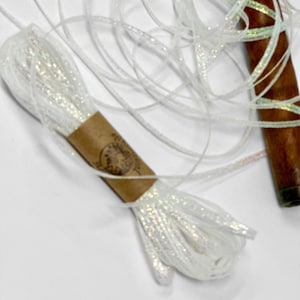 May include: A spool of white, glittery ribbon with a brown paper label. The ribbon is loosely coiled, and the glitter catches the light. A wooden object is visible in the background. This ribbon is suitable for crafts and gift wrapping.