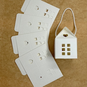 May include: Five white paper house-shaped boxes with cut-out windows and hearts. The boxes are folded flat and ready to be assembled. There is a sixth box that is already assembled and has a string attached to the top.
