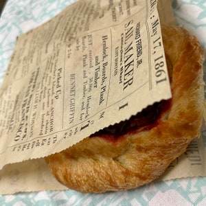 May include: A flaky pastry with a red filling wrapped in a brown paper bag with text from an old newspaper advertisement. The text reads "Richard Friend, Jr. Sail Maker Cunningham's Wharf East Boston Hemlock, Boards, Plank and Timber. Just received 30,000 Hemlock Boards, Plank and Timber, and for sale by 2-1w Bennet Griffin"