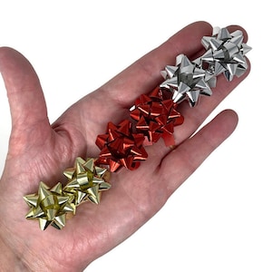 May include: Five small gift bows in different colors. The bows are red, gold, silver, and two gold bows. The bows are arranged in a row on a hand.