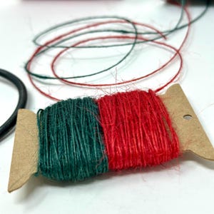 May include: Two spools of twine, one green and one red, are wrapped around a cardboard spool holder. The twine is made of natural fibers and has a rustic look.