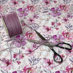 May include: A spool of purple velvet ribbon with a pair of silver scissors on a pink floral patterned background.