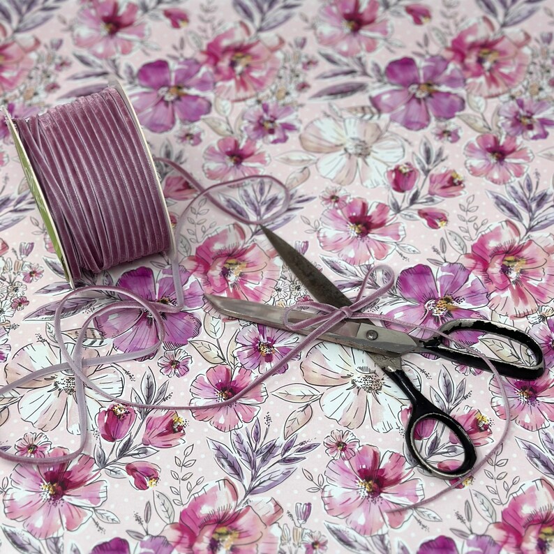 May include: A spool of purple velvet ribbon with a pair of silver scissors on a pink floral patterned background.