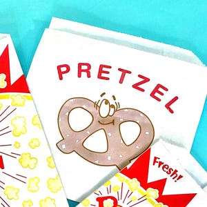 May include: White paper bags with red text that says "Pretzel" and a cartoon pretzel with a smiling face. One bag has a red and white design with the word "Fresh!" on it.