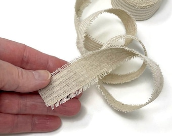 May include: A roll of natural-colored, frayed cotton ribbon with a subtle striped pattern. The ribbon is approximately 1 inch wide and has a raw, unfinished edge.