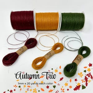May include: An image of three spools of twine in burgundy, gold, and green, with matching bundles below. The text "Autumn Trio" is displayed, along with "1mm x 20 yards each color". Decorative fall leaves are scattered around the twine.