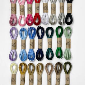 May include: A collection of colorful satin ribbons in various shades, including pink, red, blue, green, and gold. Each ribbon is neatly rolled and secured with a brown paper band, creating a visually appealing display. The ribbons are arranged on a white surface.