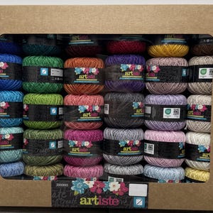 May include: A cardboard box filled with spools of colorful yarn. The yarn is in various shades, including blue, green, orange, purple, pink, gray, and yellow. Each spool is labeled with the brand name "artiste". The box has a rectangular window.