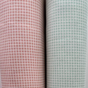 May include: Two rolls of fabric, one in a light coral color and the other in a light green, both with a small gingham check pattern. The fabric is tightly rolled, showing the texture and pattern clearly. The rolls are side by side.