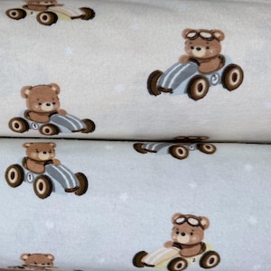 May include: Two rolls of fabric featuring a repeating pattern of brown teddy bears wearing goggles and driving vintage-style race cars. The top roll is off-white, and the bottom roll is light blue, both decorated with small white star accents.