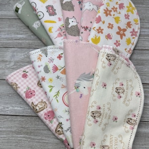 May include: A collection of baby burp cloths with various patterns. Designs include hedgehogs, bunnies, floral arrangements, and a unicorn. The cloths are in shades of pink, white, and green, with scalloped edges.