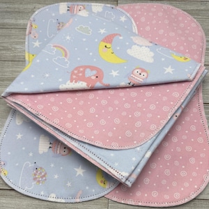 May include: A stack of baby burp cloths. One cloth is light blue with a pattern of a rainbow, moon, stars, clouds, and an elephant. The other cloth is pink with a white swirl pattern. The edges are stitched with a contrasting thread.