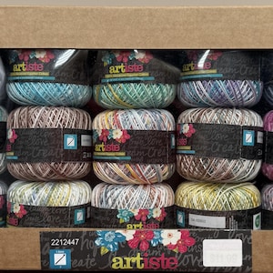 May include: A cardboard box filled with spools of hand-dyed yarn in various colors, including blue, green, brown, pink, and yellow. Each spool is labeled with the brand name "artiste" and floral accents. The box has a circular sticker that says "hand dyed".
