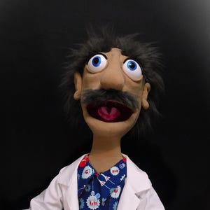 Könnte beinhalten: Eine Puppe eines Arztes mit einem großen Schnurrbart, die einen weißen Laborkittel, ein blau gemustertes Hemd, eine rote Hose und schwarze Schuhe trägt. Die Puppe hat große blaue Augen und einen offenen Mund.