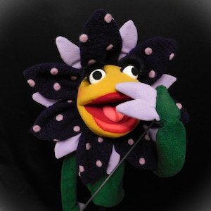May include: A flower puppet with a yellow face, red lips, and a purple tongue. The flower has dark blue petals with pink polka dots and light purple accents. It has a green stem against a black background. A fun, colourful puppet.