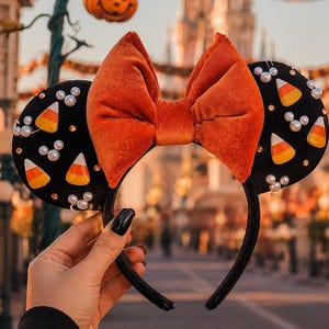 May include: A black headband featuring two circular ears decorated with white pearl beads and candy corn shaped accents. A large, plush orange velvet bow is centered between the ears. The headband has a soft black fabric covering the entire frame.